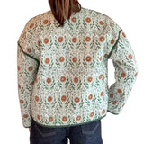 A person viewed from behind wearing a quilted jacket with a green and orange floral pattern on a cream background, paired with dark blue jeans, standing against a white paneled wall.