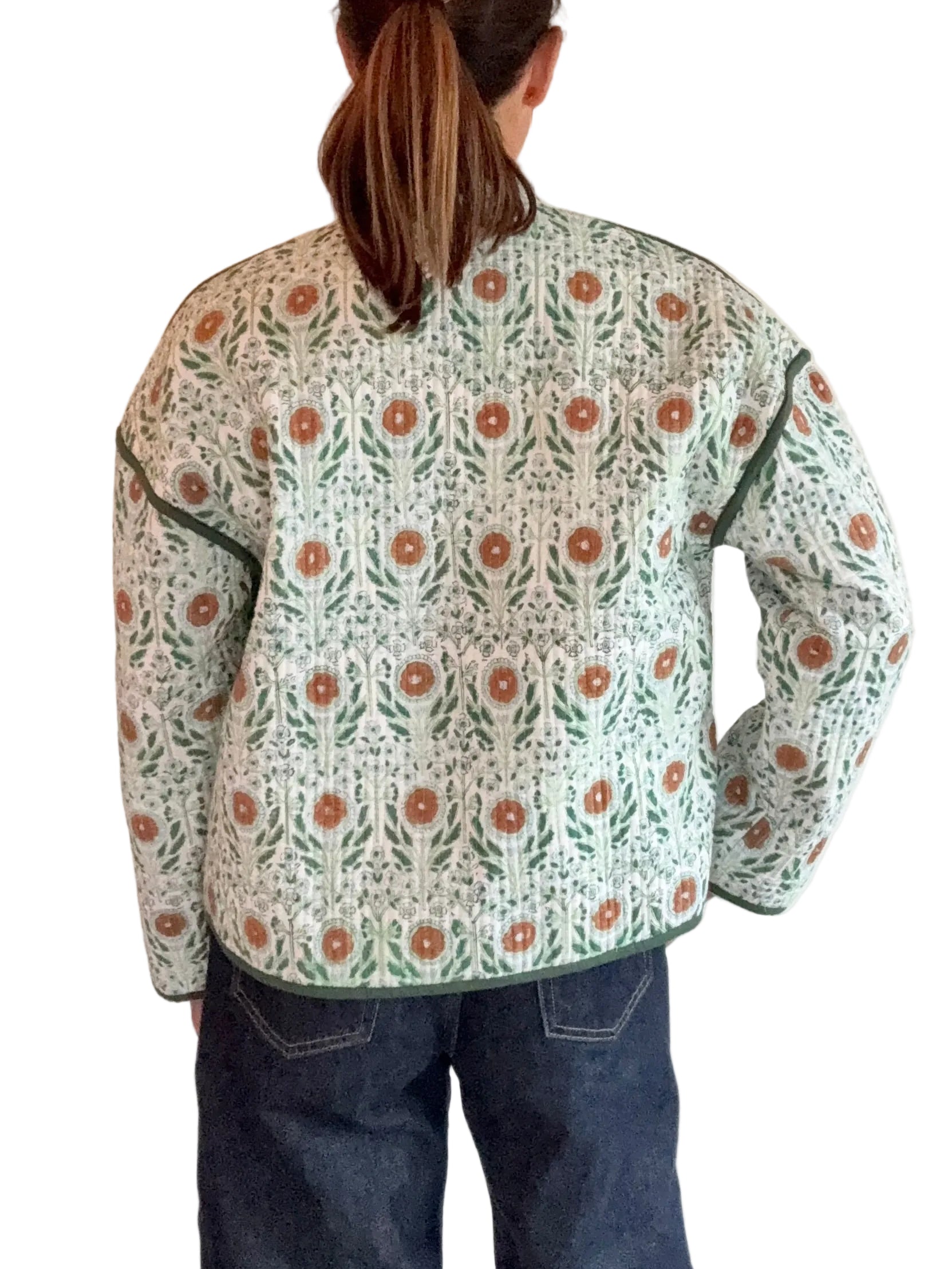 A person viewed from behind wearing a quilted jacket with a green and orange floral pattern on a cream background, paired with dark blue jeans, standing against a white paneled wall.