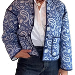 A person wearing a blue and white paisley patterned jacket over a white button-down shirt and dark jeans, standing against a white paneled wall.