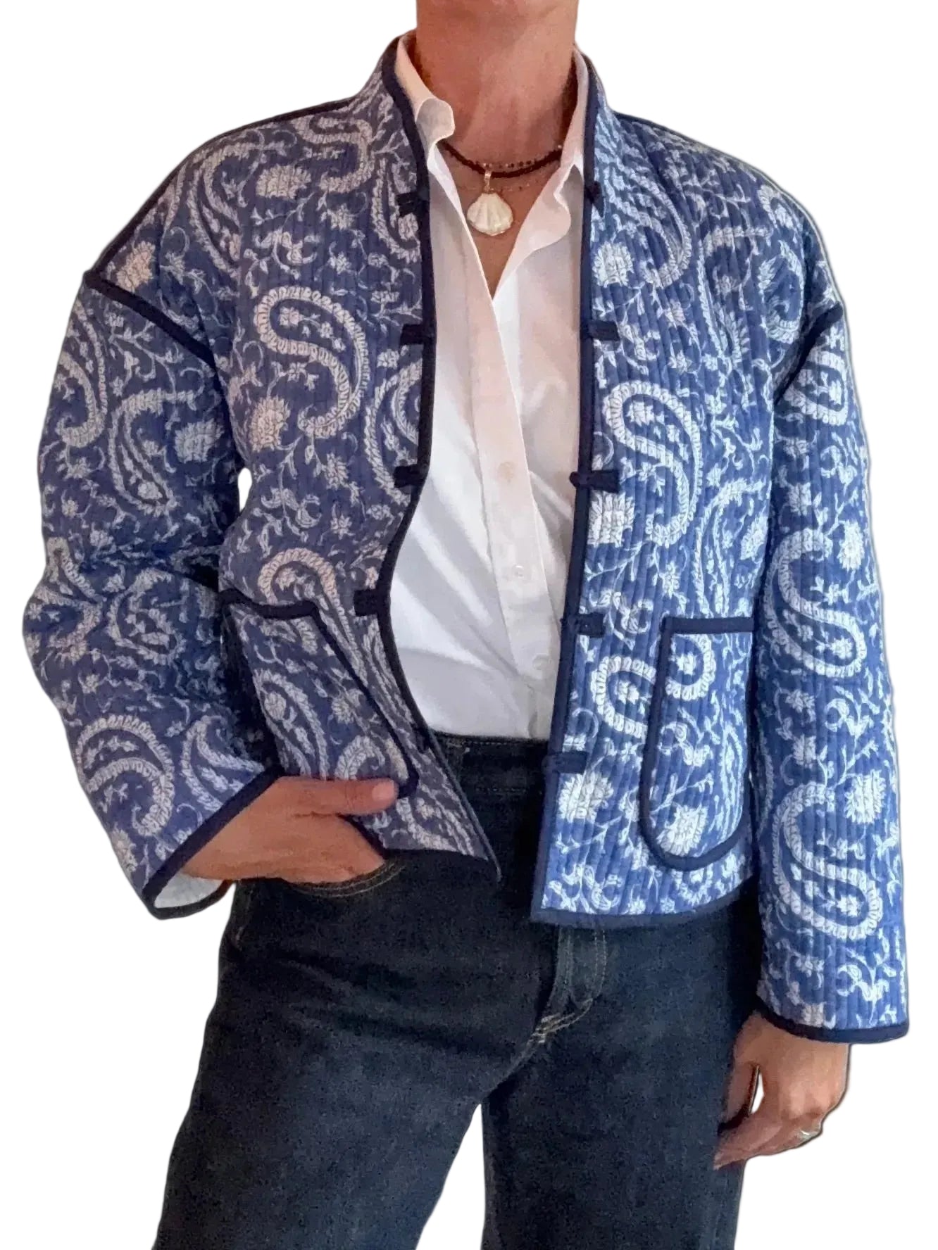 A person wearing a blue and white paisley patterned jacket over a white button-down shirt and dark jeans, standing against a white paneled wall.